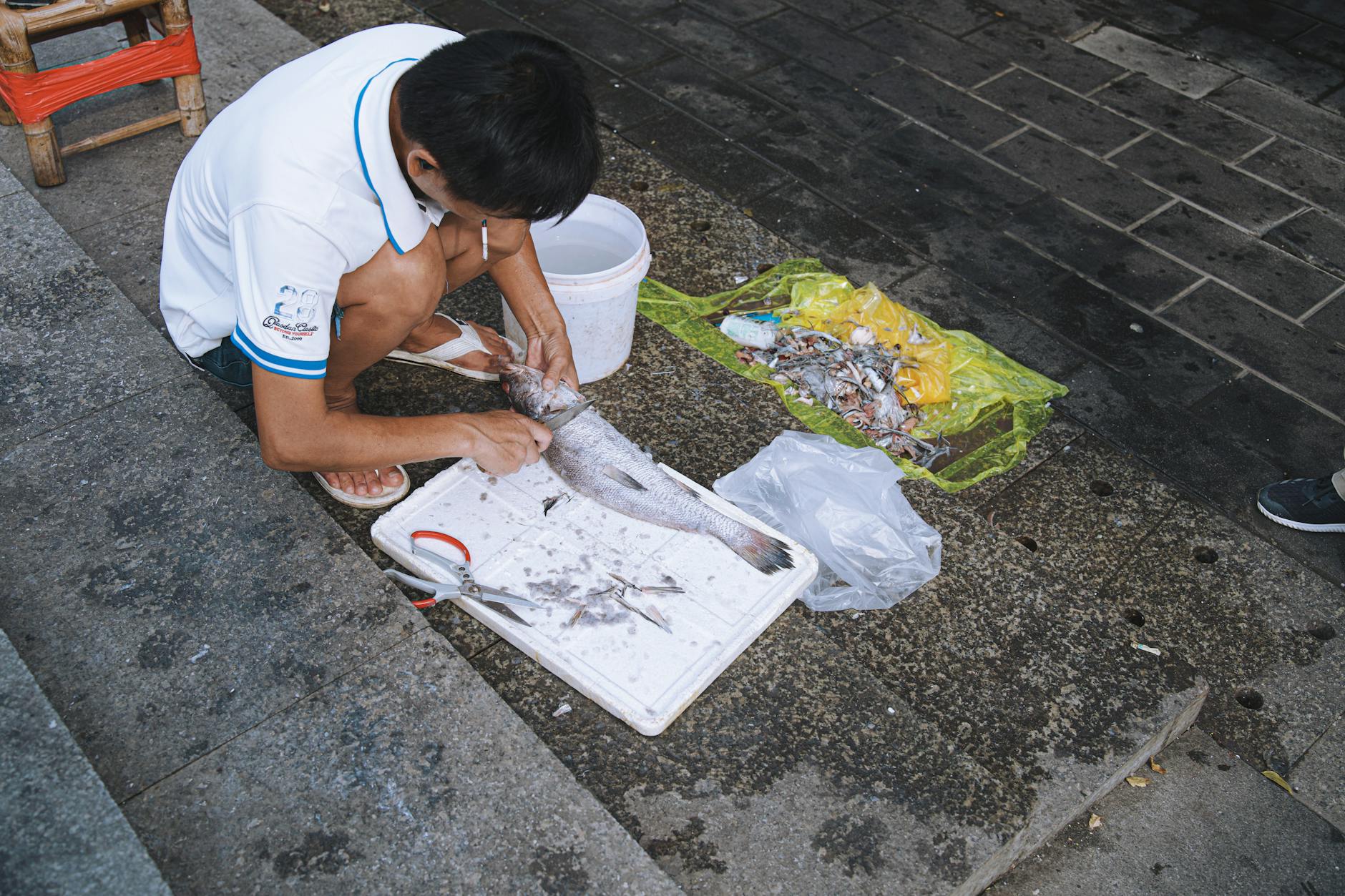 A man crouches on steps scaling fish, surrounded by urban textures and waste.