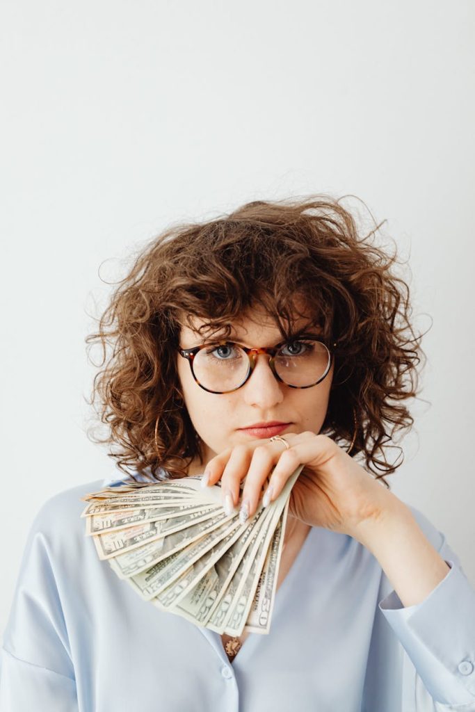 Budget-Friendly Salvage: 5 Essential Tips for Profitable Success Caucasian woman with curly hair and glasses holding US dollars confidently indoors.