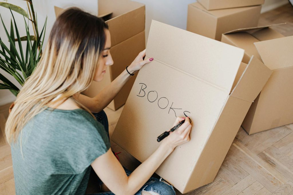 Home Move Hacks: Effortless Tips to Declutter Before Your Move Woman writing on cardboard box while packing books, preparing for moving day at home.