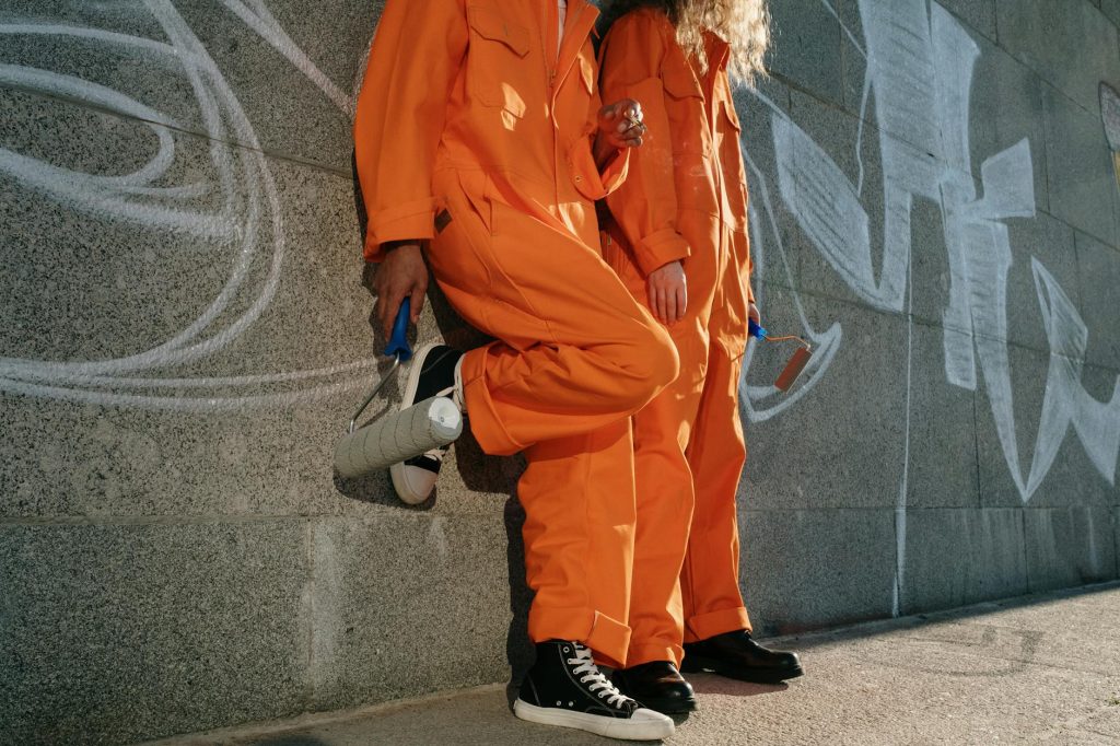 Industrial Removals: Best Tips for Choosing the Right Team Two people in orange uniforms with paint rollers leaning on a graffiti-covered wall outdoors.