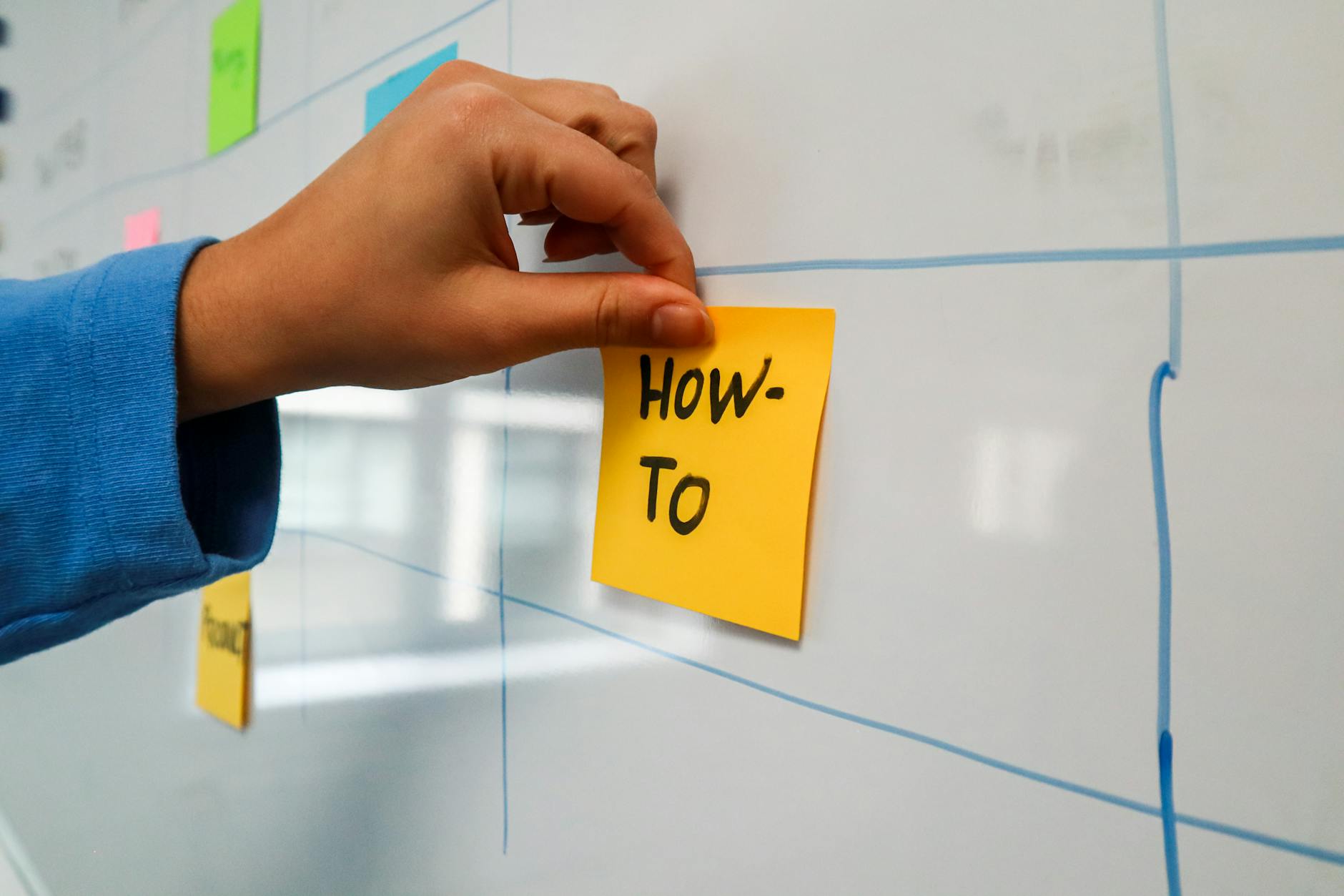 Relocate Workshop Effortlessly: Maintain Productivity Close-up of a hand placing a yellow 'How-To' sticky note on a whiteboard for planning.