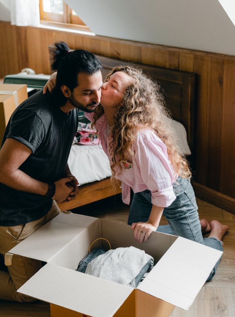 New Home Moving Tips: Effortless Ways to Settle In Cheerful female in casual wear kissing boyfriend in cheek and sitting on floor while moving house and unpacking boxes
