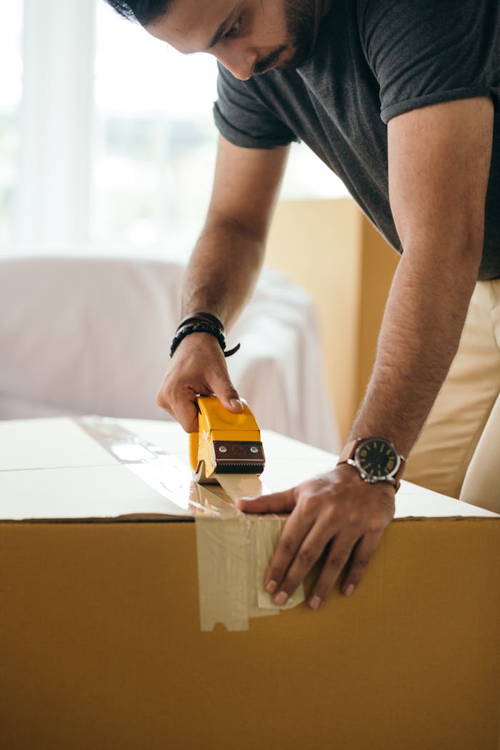 Home Side view of concentrated crop ethnic male sticking cardboard box with tape during preparing for moving in house at daytime