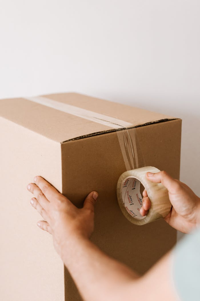 About Crop anonymous male using tape to seal packed cardboard box against white wall while moving into new place