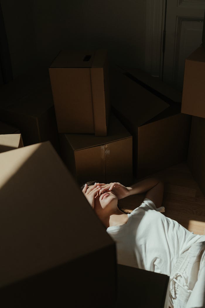 Menus A woman lies on the floor surrounded by cardboard boxes in a dim sunlit room, expressing fatigue.